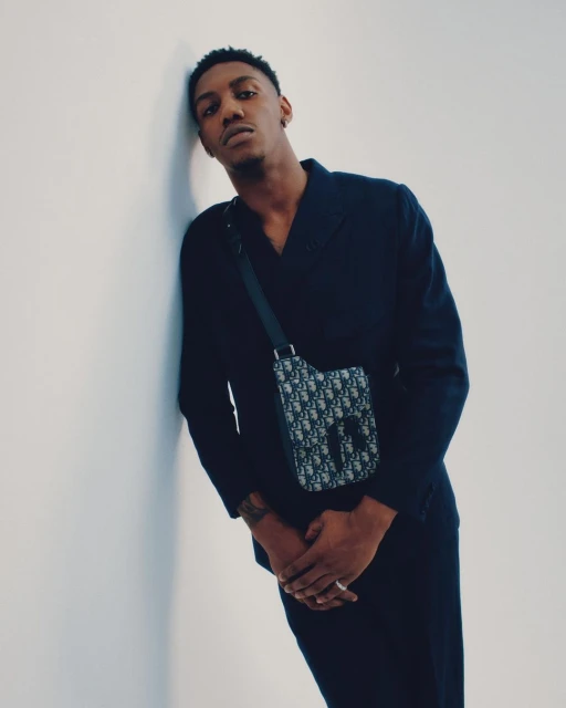 Portrait of a young adult Black male in dark clothing leaning against a white wall with a branded crossbody bag.