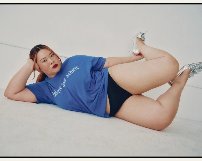 A plus-size woman in a blue t-shirt and black underwear reclines on a white surface wearing high heels, indoors, with red lipstick.