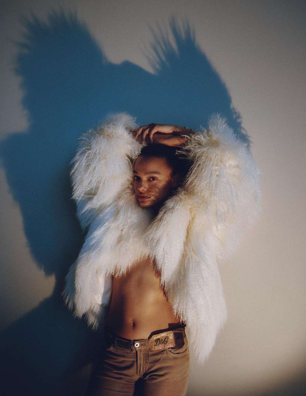 Adult with pale skin, wearing a white shaggy jacket and tan pants. Posing indoors against a white wall with a light blue shadow.
