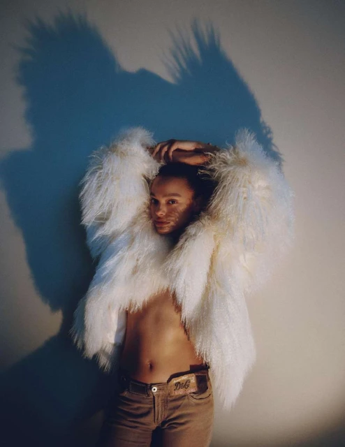 Adult with pale skin, wearing a white shaggy jacket and tan pants. Posing indoors against a white wall with a light blue shadow.