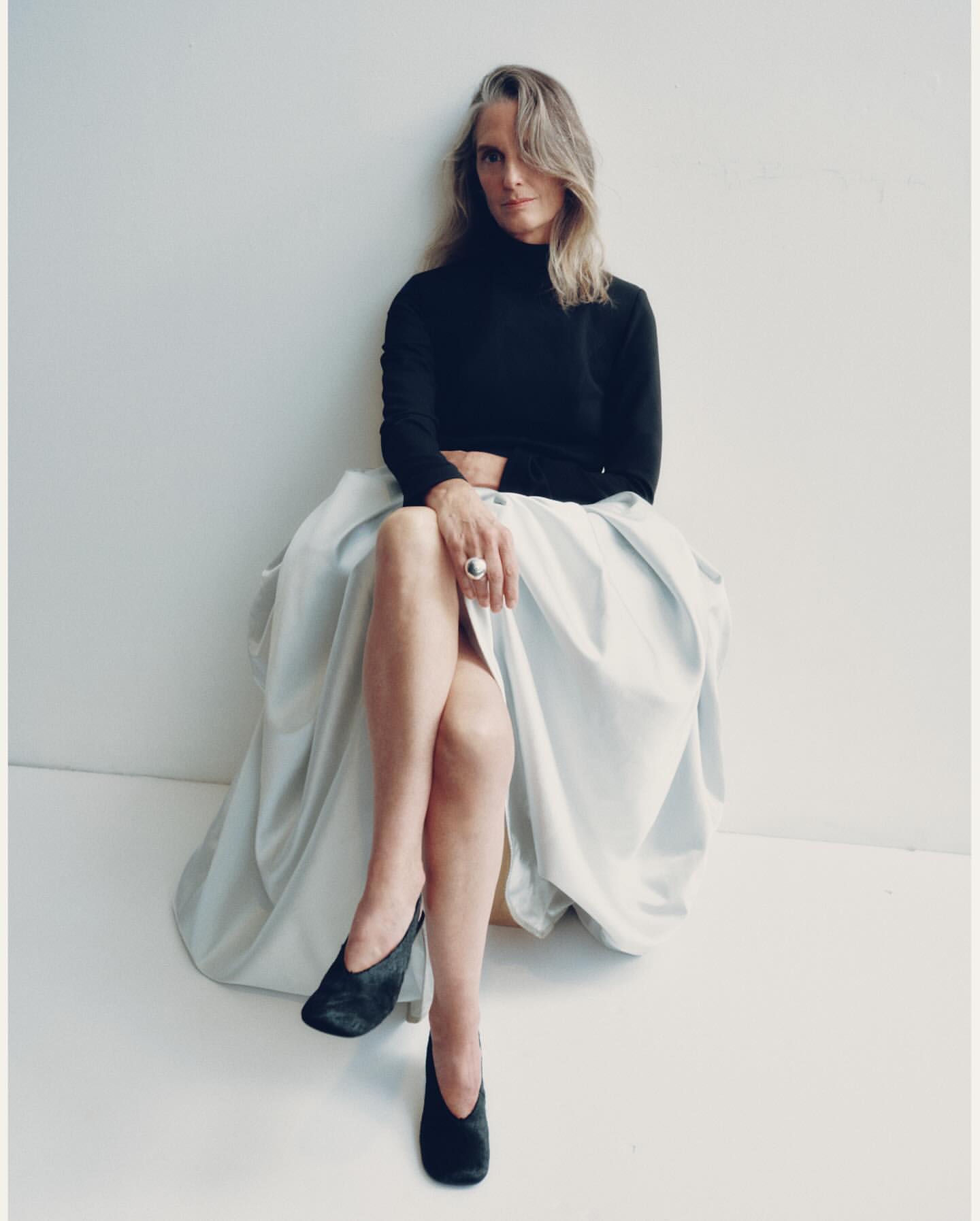Mature woman with long blonde hair wearing a black turtleneck and voluminous pale skirt, seated against a white background.