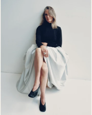 Mature woman with long blonde hair wearing a black turtleneck and voluminous pale skirt, seated against a white background.