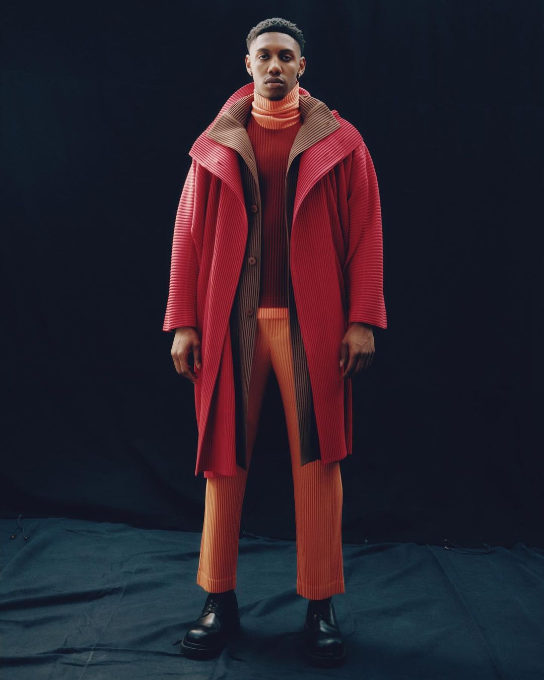 Full-length portrait of a Black man wearing a red coat and orange ribbed turtleneck against a dark seamless backdrop.