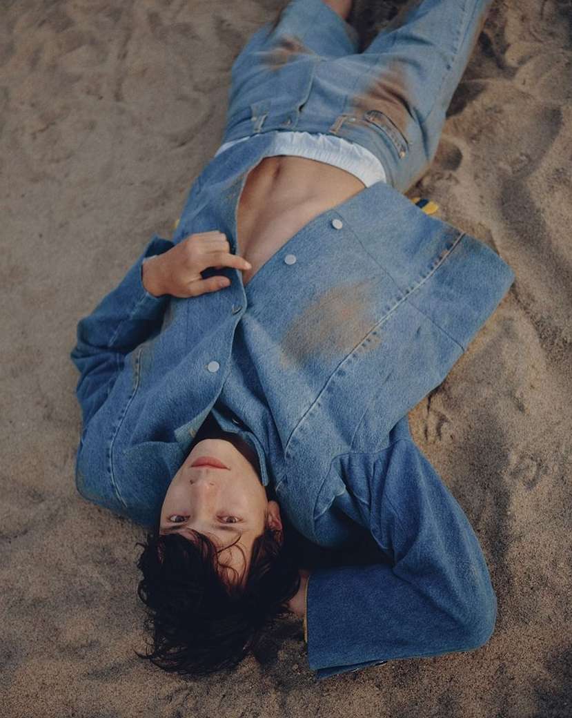 Person in denim jacket and jeans laying in the sand with an open shirt, showing their torso, hair is dark and messy. 