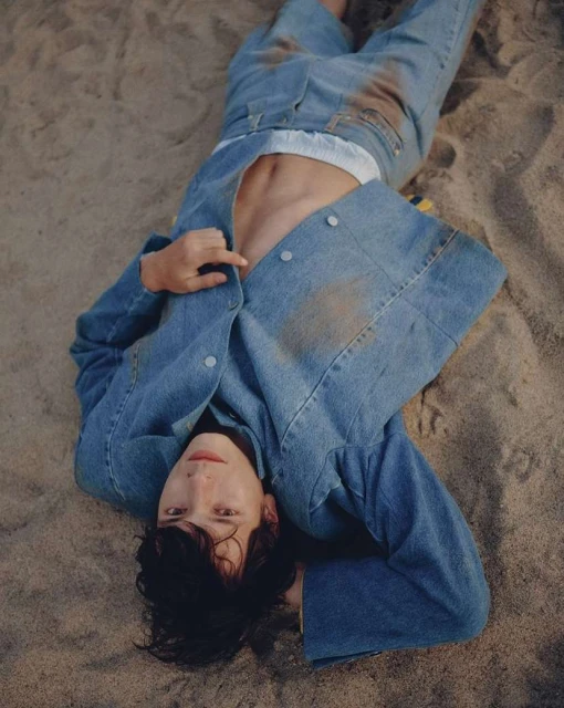 Person in denim jacket and jeans laying in the sand with an open shirt, showing their torso, hair is dark and messy. 