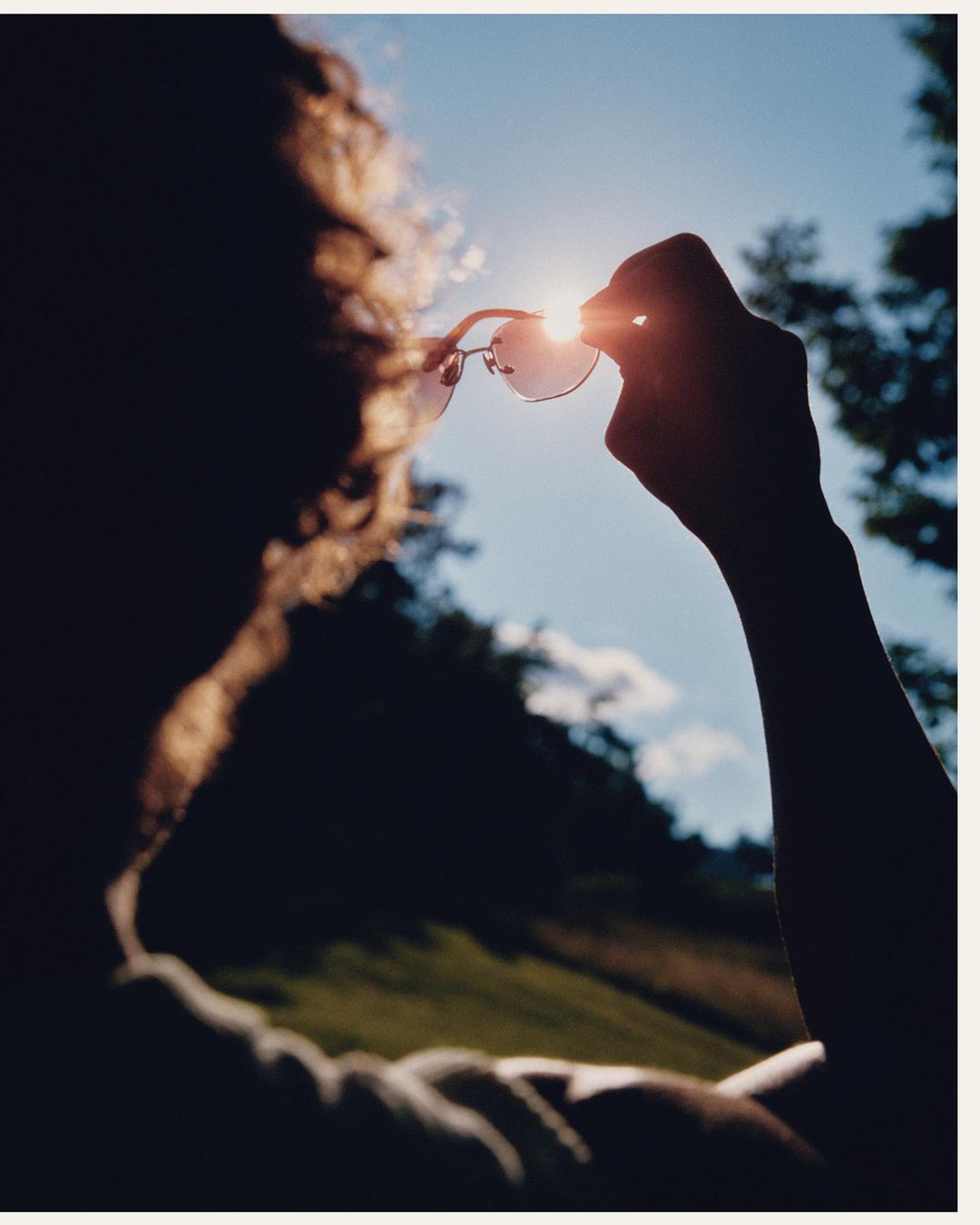 Person holding sunglasses up to the sun with silhouetted head and hand with a view of trees and sky in the background.