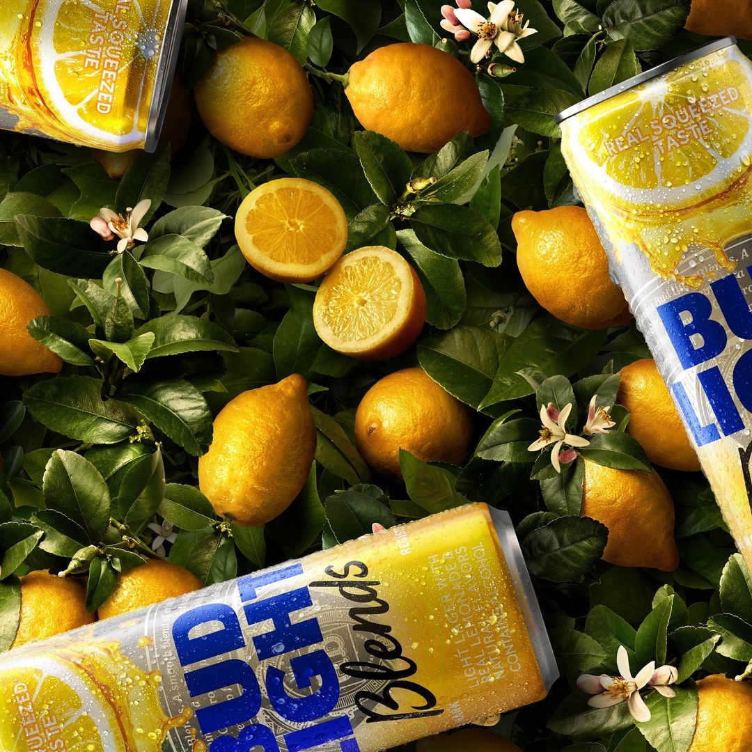 Overhead shot of lemons, lemon slices, lemon blossoms and Bud Light Blends cans scattered across a leafy surface.