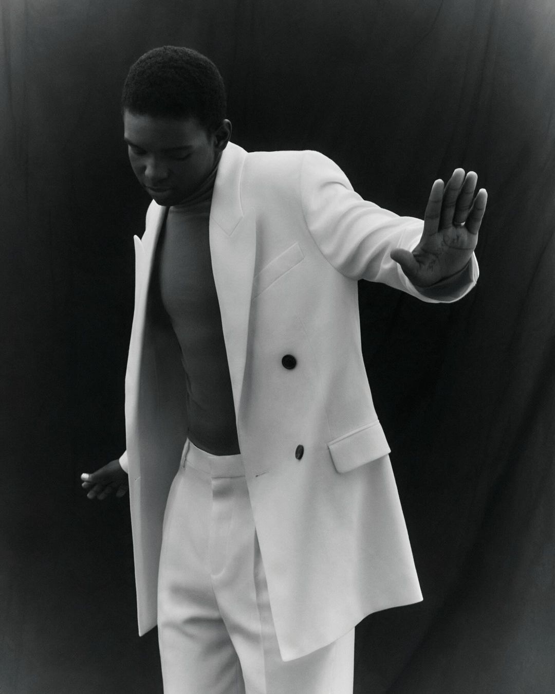 Young black man in a white suit jacket and trousers, posing in front of a dark background, his hand slightly raised.
