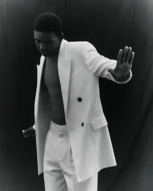 Young black man in a white suit jacket and trousers, posing in front of a dark background, his hand slightly raised.