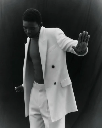 Young black man in a white suit jacket and trousers, posing in front of a dark background, his hand slightly raised.