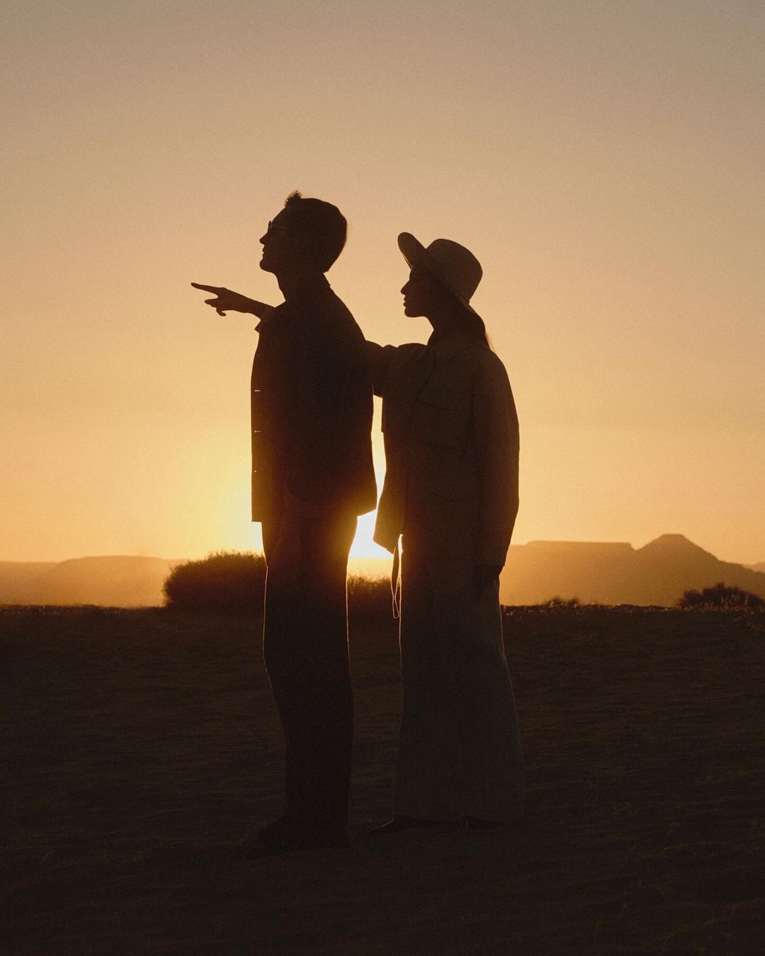 Silhouette of a man and woman standing in a field at sunset, with mountains visible in the background, creating a warm and golden atmosphere.