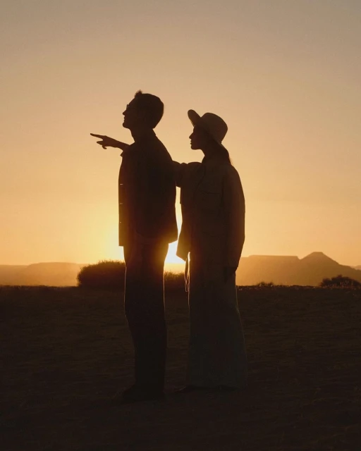 Silhouette of a man and woman standing in a field at sunset, with mountains visible in the background, creating a warm and golden atmosphere.