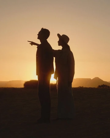 Silhouette of a man and woman standing in a field at sunset, with mountains visible in the background, creating a warm and golden atmosphere.