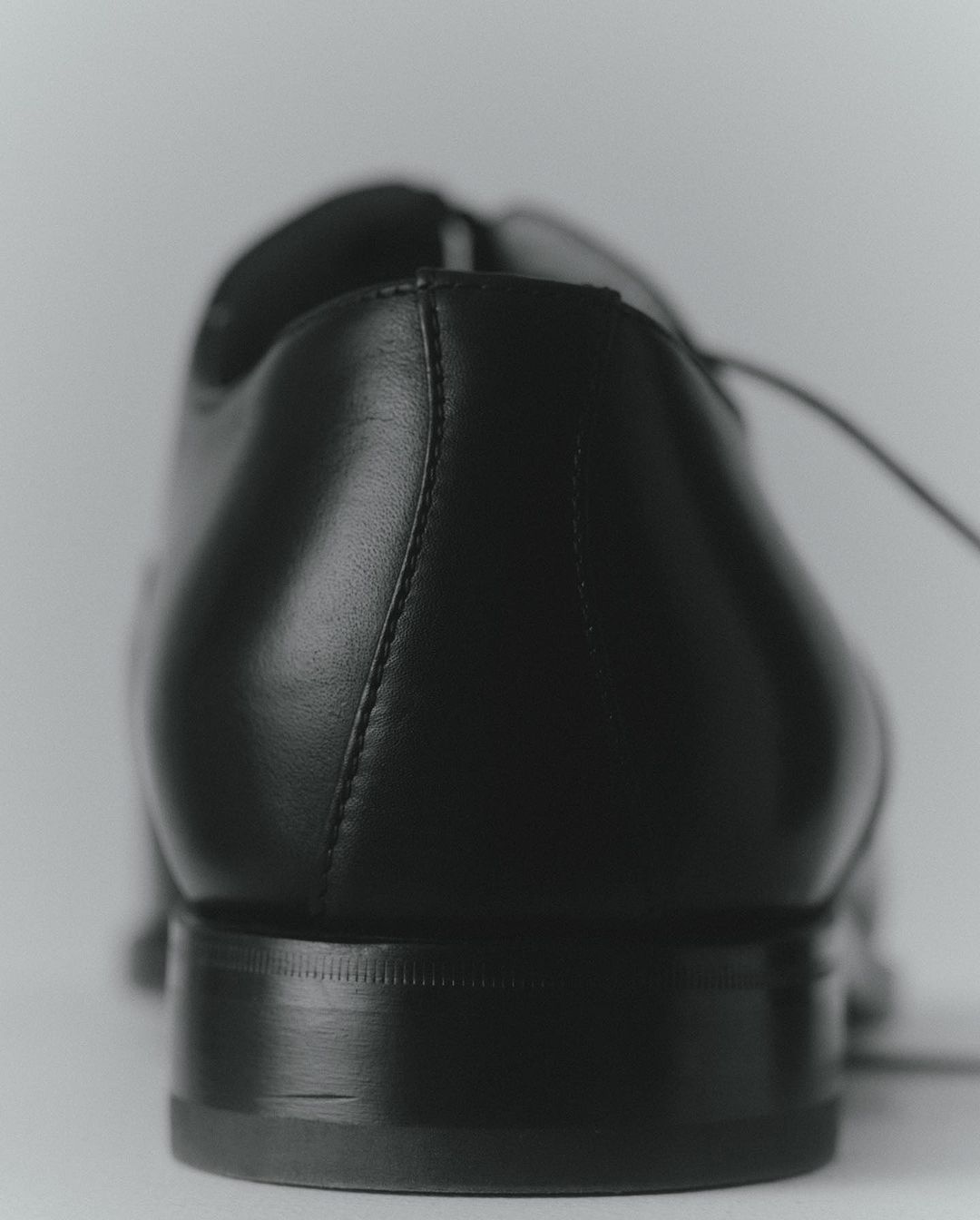 Close-up of a black leather dress shoe showing the heel, counter, and laces, set against a neutral, light-colored background.