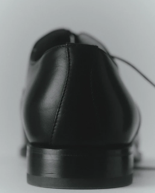 Close-up of a black leather dress shoe showing the heel, counter, and laces, set against a neutral, light-colored background.
