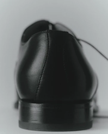 Close-up of a black leather dress shoe showing the heel, counter, and laces, set against a neutral, light-colored background.