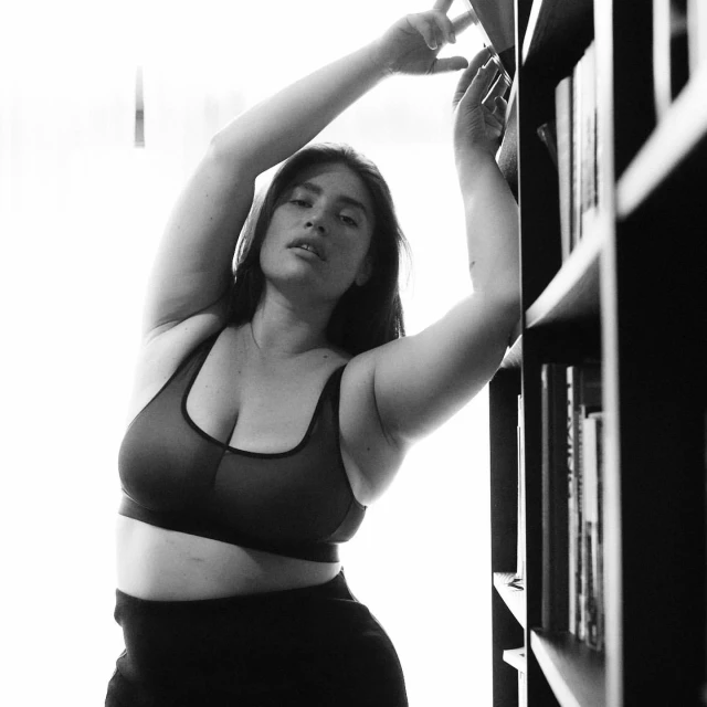 A woman in a sports bra stands in front of bookshelves in a monochrome photo. Arms are raised above her head, stretching.