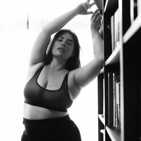 A woman in a sports bra stands in front of bookshelves in a monochrome photo. Arms are raised above her head, stretching.