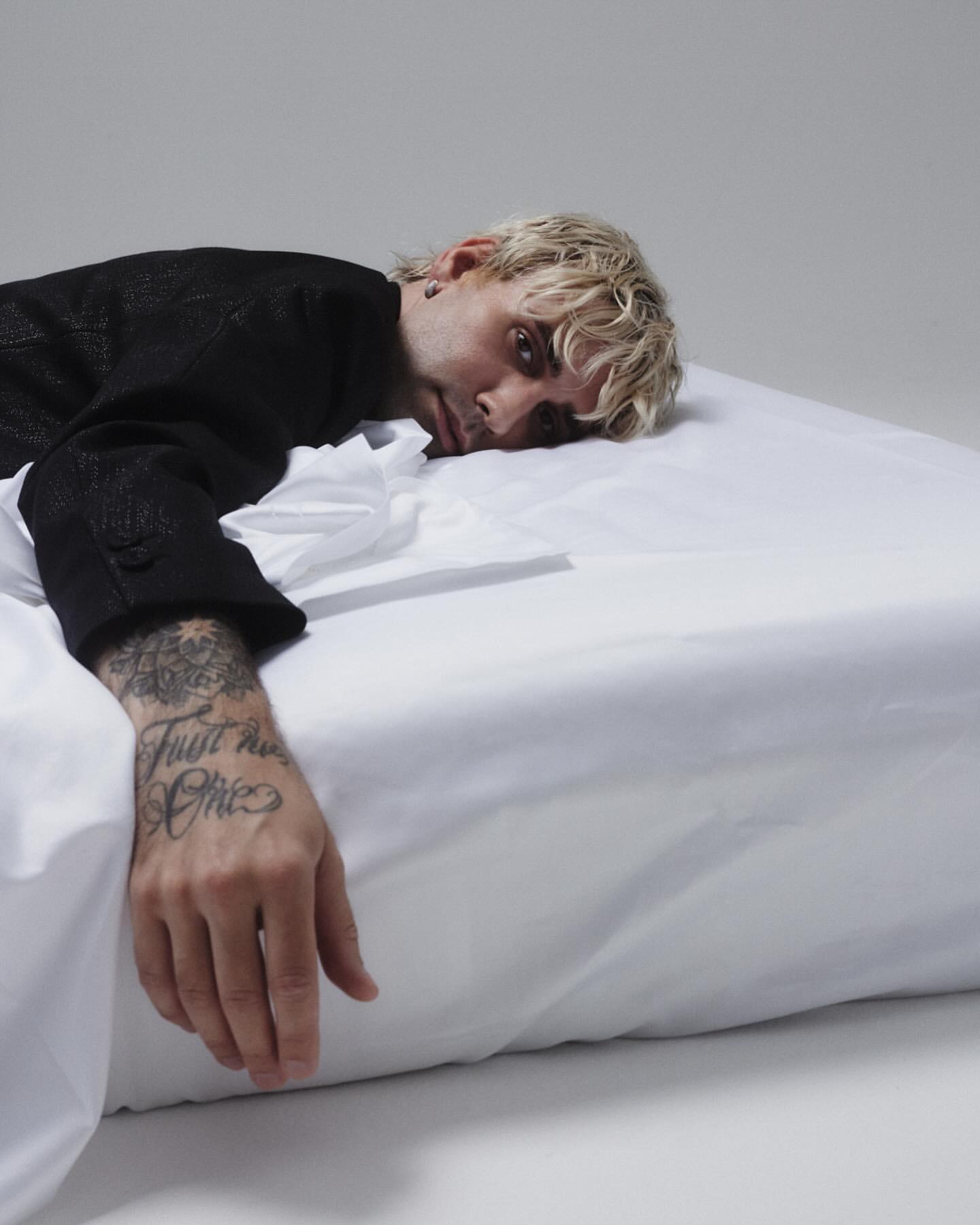 Man with blond hair and a black jacket lies on a white bed, facing the camera in a relaxed pose against a simple grey background.