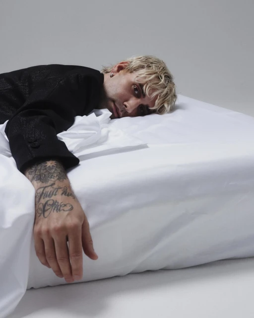 Man with blond hair and a black jacket lies on a white bed, facing the camera in a relaxed pose against a simple grey background.