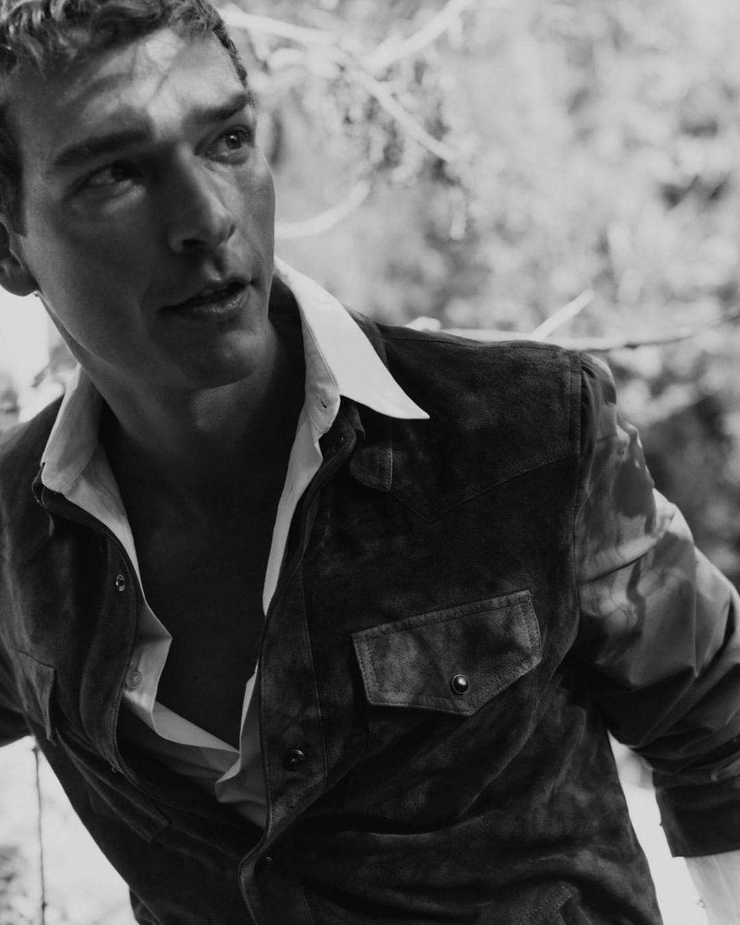 Black and white portrait of a man wearing a suede jacket and unbuttoned white shirt, captured outdoors with natural lighting and a forest background.