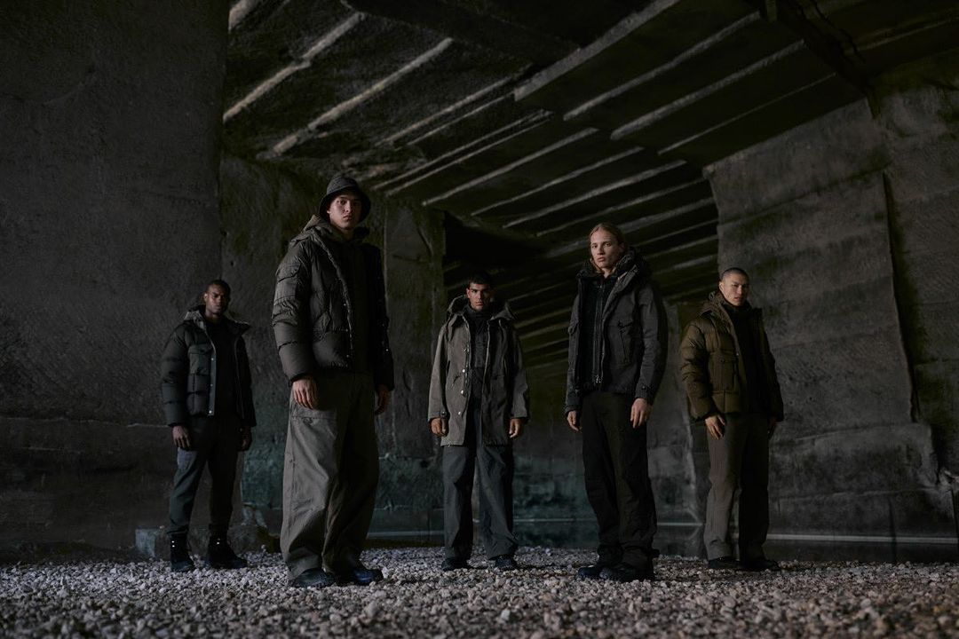 Five people standing in dark indoor setting, wearing puffer jackets. The setting looks like a stone quarry or mine.