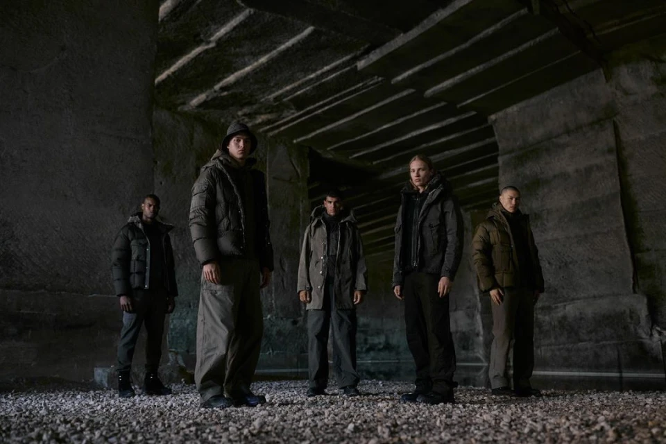 Five people standing in dark indoor setting, wearing puffer jackets. The setting looks like a stone quarry or mine.