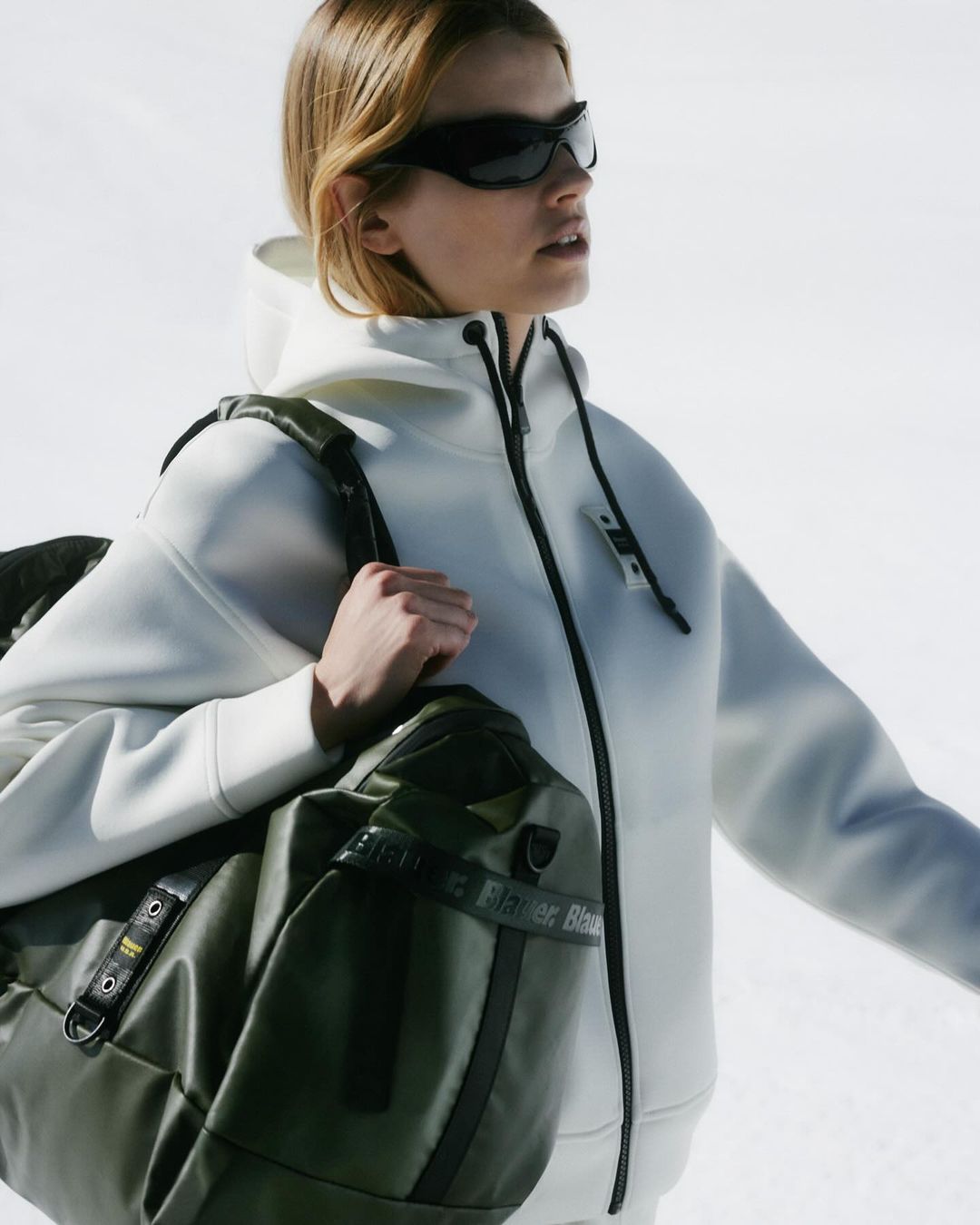 Woman wearing sunglasses and a hoodie carrying a duffel bag. The background shows snow. Fashion, outdoor lifestyle and sport.