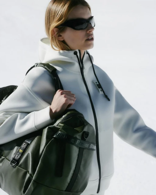 Woman wearing sunglasses and a hoodie carrying a duffel bag. The background shows snow. Fashion, outdoor lifestyle and sport.
