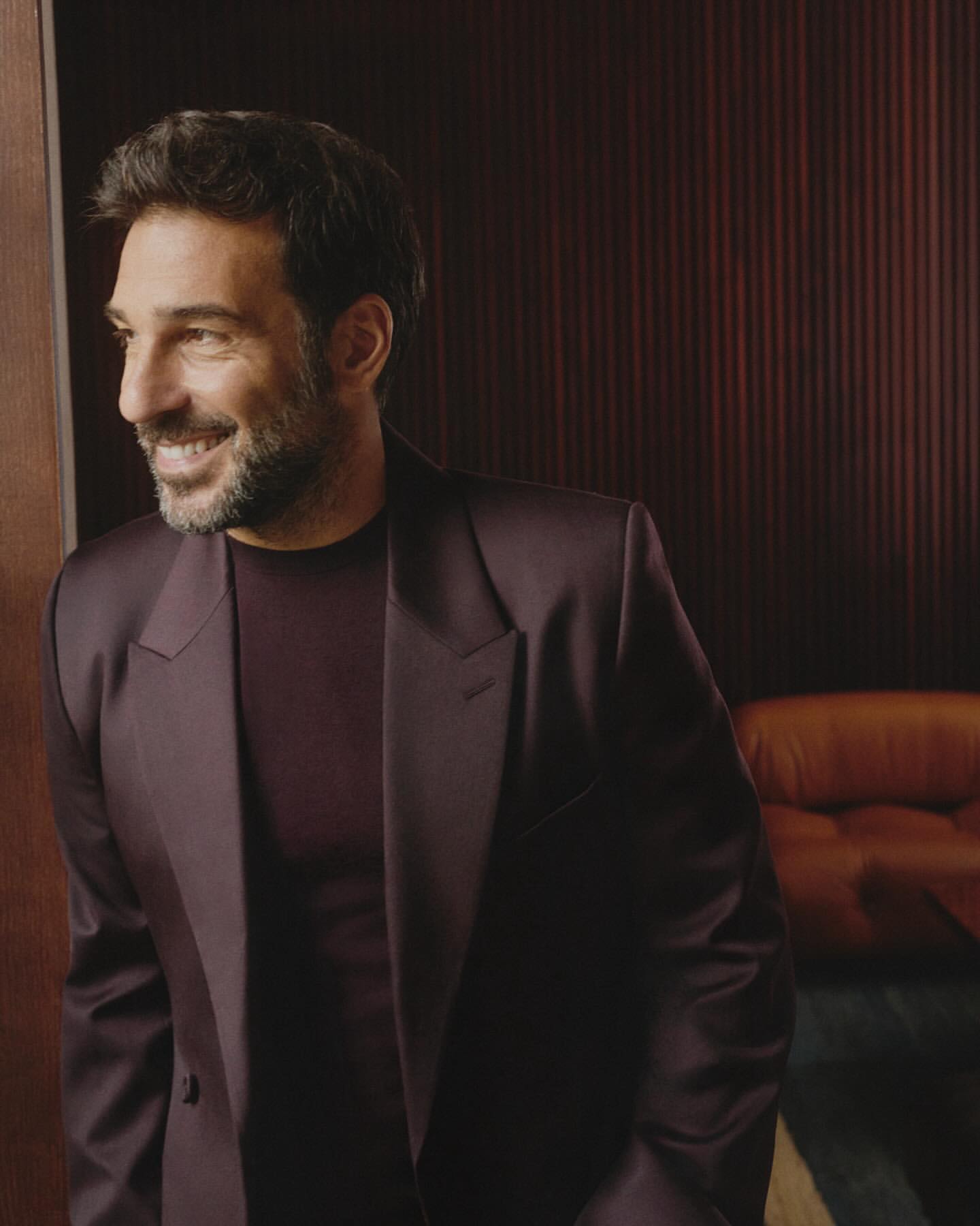 Adult male in a burgundy suit and shirt smiles in a close-up portrait inside with dark red paneled wall and brown couch background.