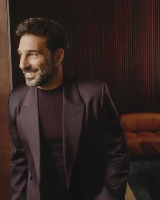 Adult male in a burgundy suit and shirt smiles in a close-up portrait inside with dark red paneled wall and brown couch background.