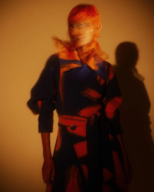 An adult fashion model poses in a blue-and-red patterned belted coat with blurred motion, lit by warm-toned lighting in a studio setting.