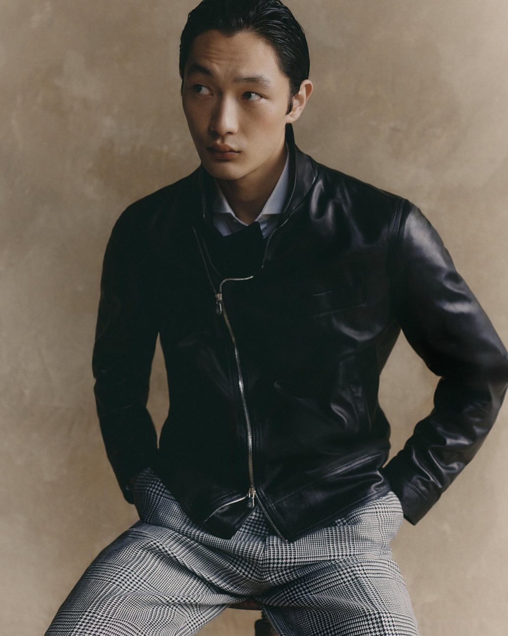 Portrait of an adult male in a black leather jacket and plaid pants, posing indoors against a neutral backdrop in soft lighting.
