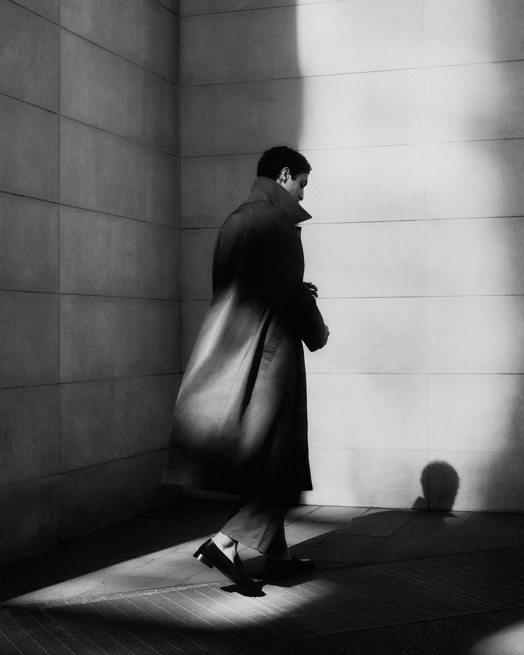 A man in a dark overcoat walks along a bright white tile wall with shadows, in a monochromatic, urban fashion scene.