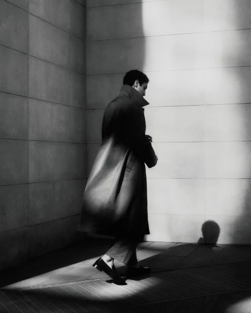 A man in a dark overcoat walks along a bright white tile wall with shadows, in a monochromatic, urban fashion scene.