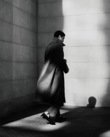 A man in a dark overcoat walks along a bright white tile wall with shadows, in a monochromatic, urban fashion scene.
