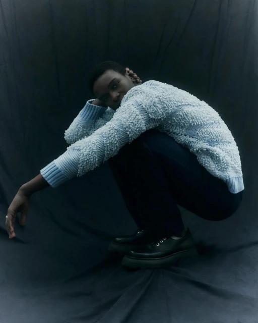 Studio shot of a Black male model crouching in a blue textured sweater, black pants, and chunky platform loafers against a dark backdrop.