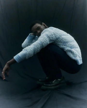 Studio shot of a Black male model crouching in a blue textured sweater, black pants, and chunky platform loafers against a dark backdrop.