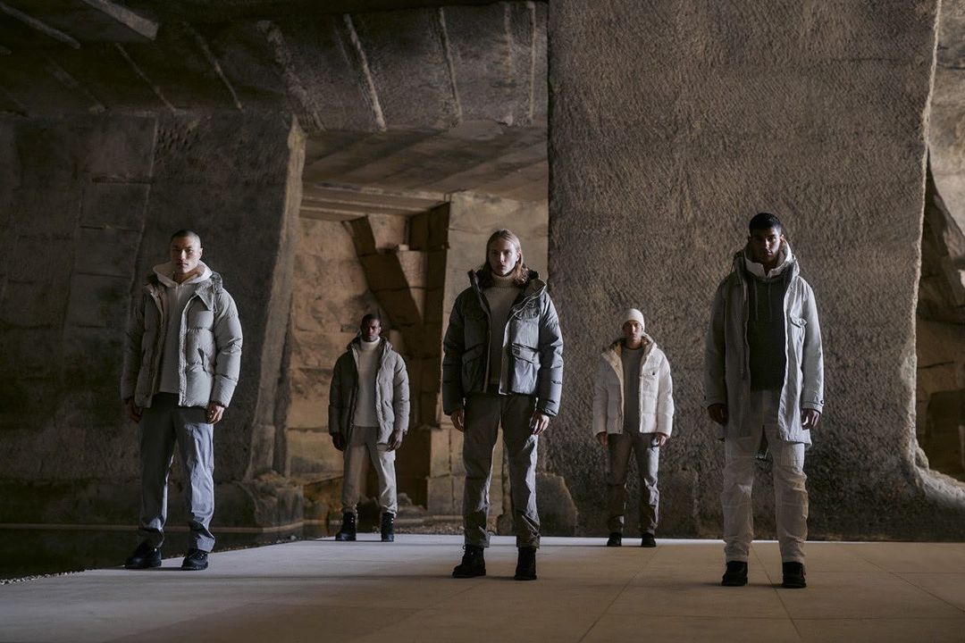 Five people stand in a stone structure, wearing winter jackets and pants, under soft lighting, creating a fashionable editorial shot.