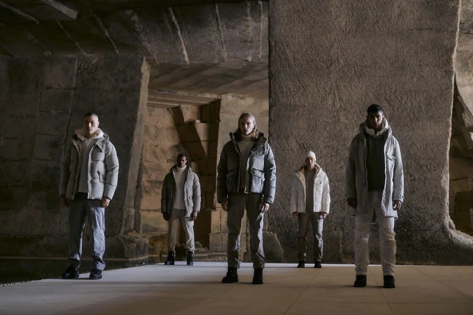 Five people stand in a stone structure, wearing winter jackets and pants, under soft lighting, creating a fashionable editorial shot.