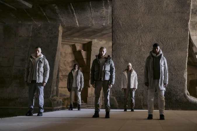 Five people stand in a stone structure, wearing winter jackets and pants, under soft lighting, creating a fashionable editorial shot.