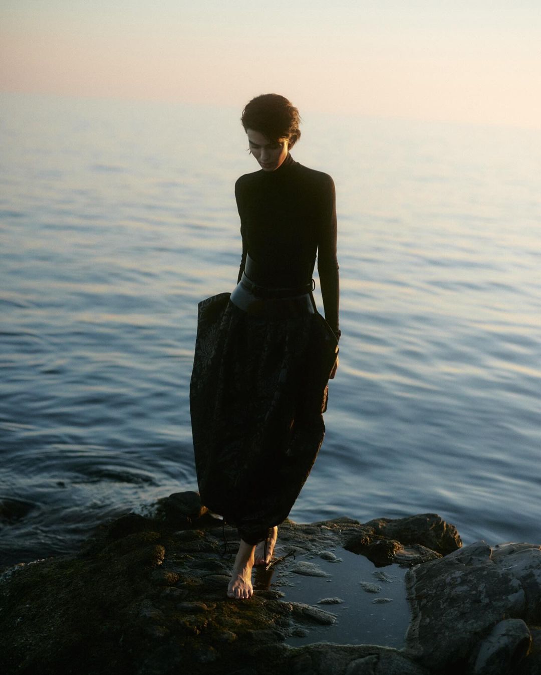 Woman in black turtleneck and dark skirt stands barefoot on rocks by the sea at dusk, gazing downward in a tranquil, fashion-editorial composition.
