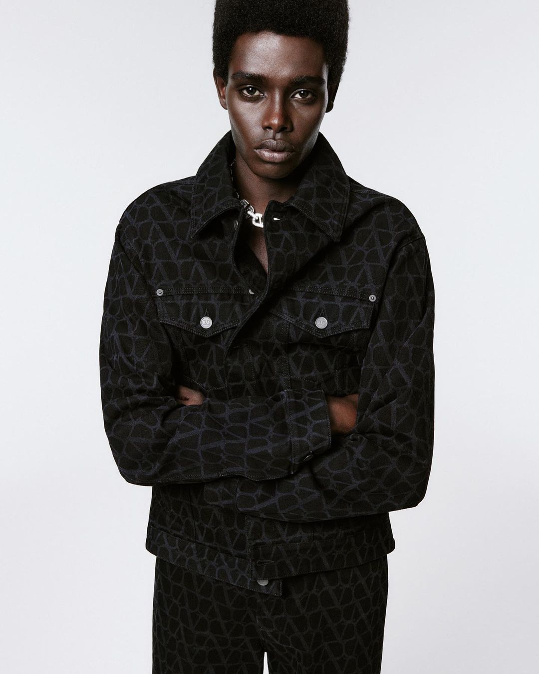Portrait of a Black man wearing a patterned denim jacket with his arms crossed against a white background in a studio setting.
