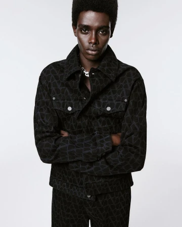Portrait of a Black man wearing a patterned denim jacket with his arms crossed against a white background in a studio setting.