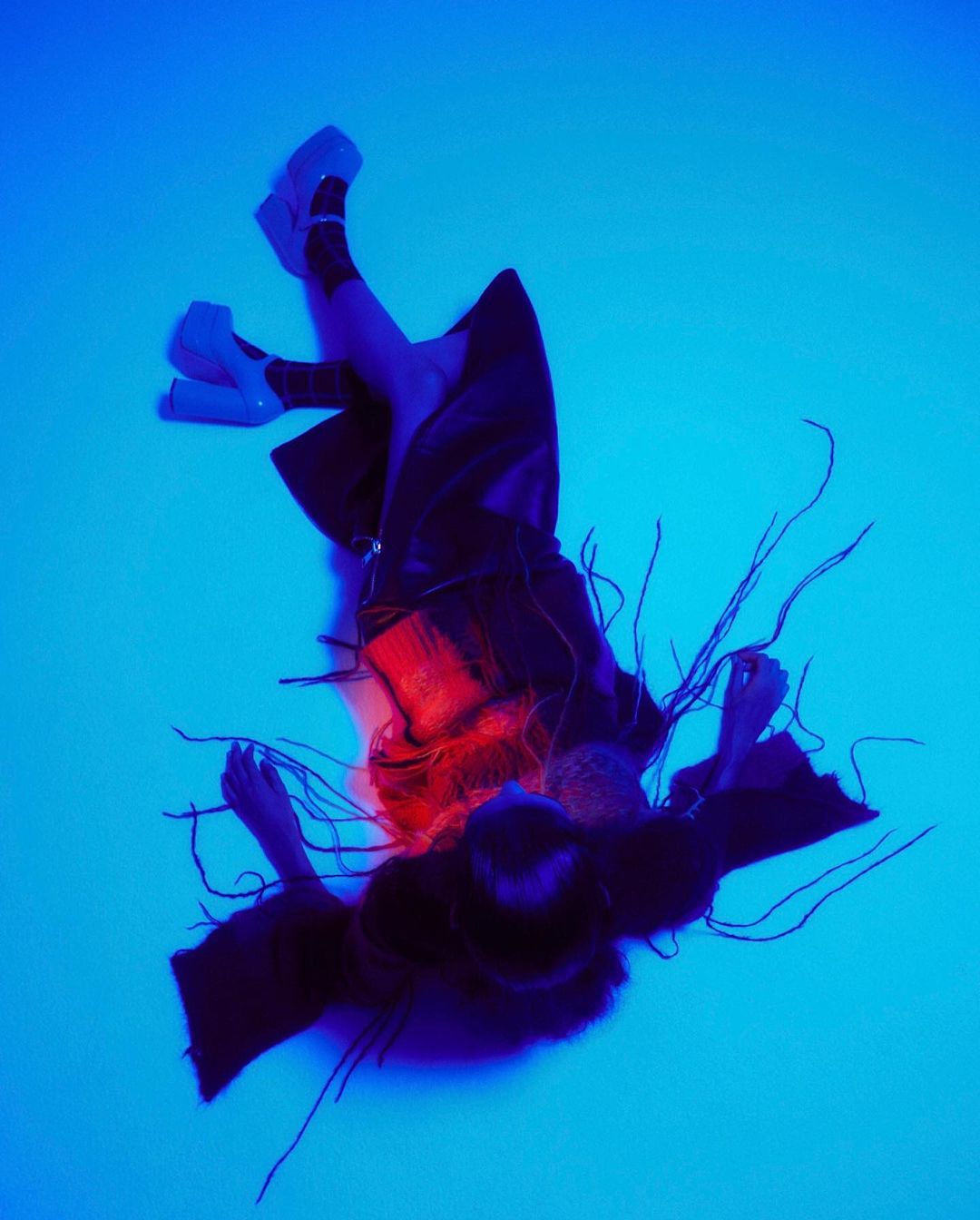 Artistic fashion photo featuring a woman in a dark outfit and platform shoes against a vibrant blue backdrop with unique red hair accents.