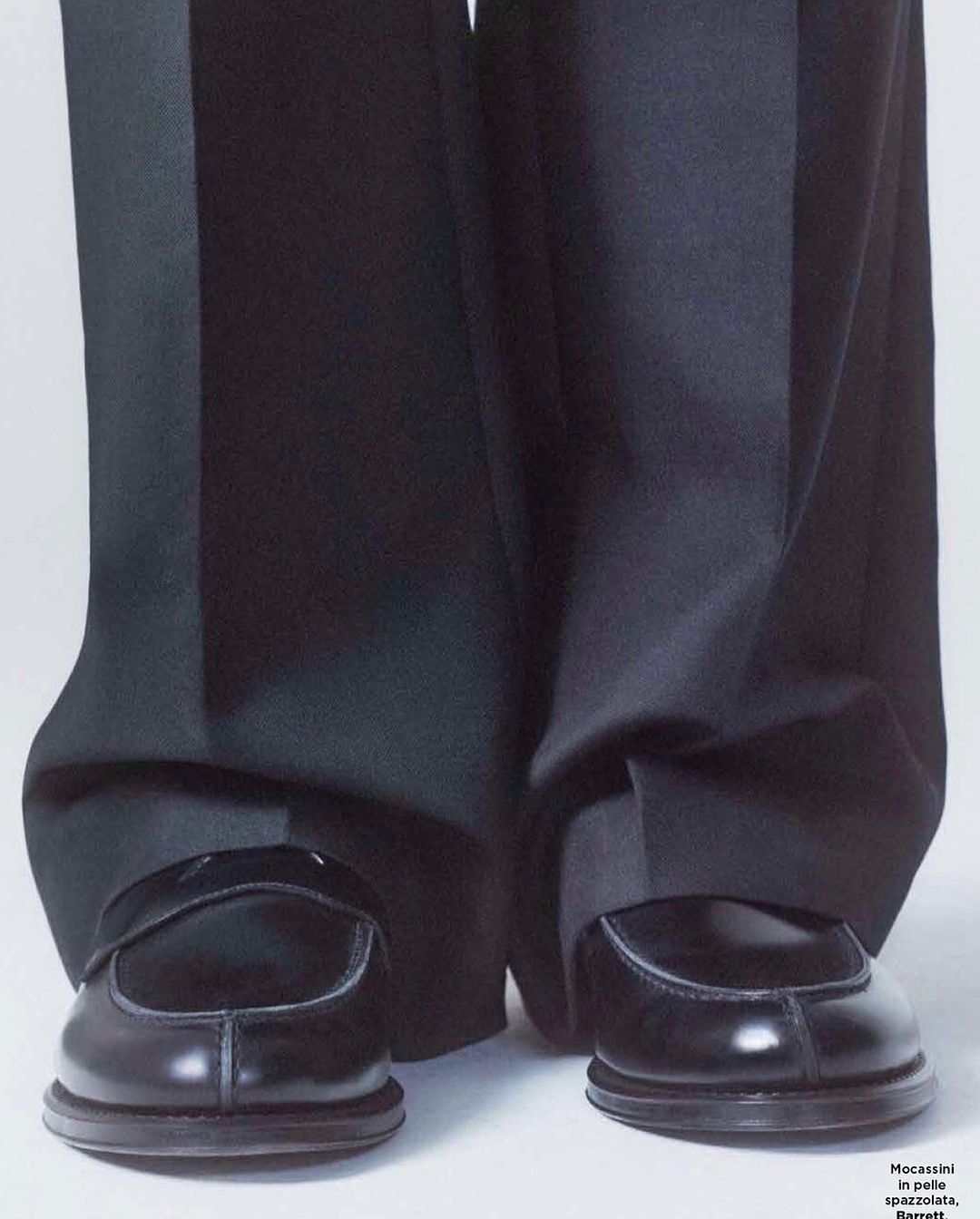 Close-up studio shot features shiny black leather penny loafer shoes and wide leg black dress pants against a white backdrop.