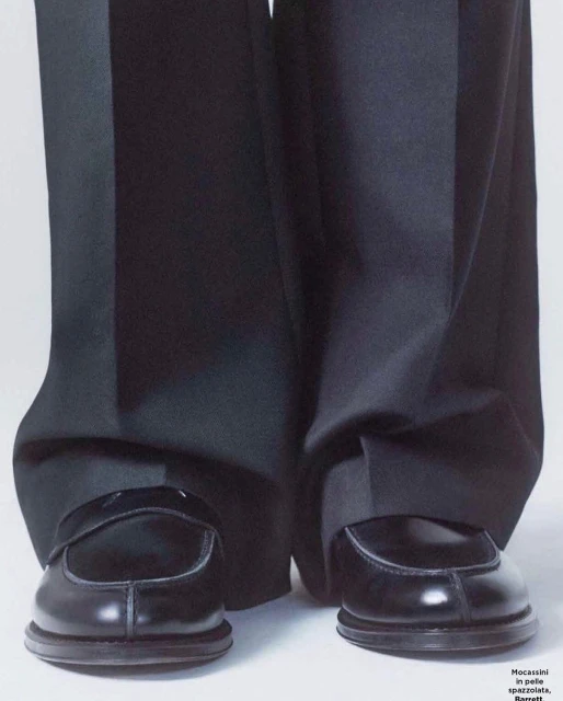 Close-up studio shot features shiny black leather penny loafer shoes and wide leg black dress pants against a white backdrop.