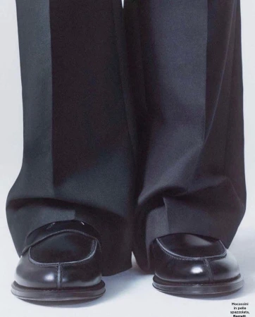 Close-up studio shot features shiny black leather penny loafer shoes and wide leg black dress pants against a white backdrop.
