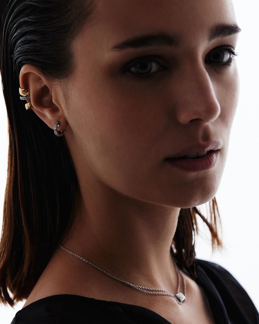 Close-up portrait of a woman with dark hair wearing earrings and a necklace against a bright white background, focusing on jewelry details.