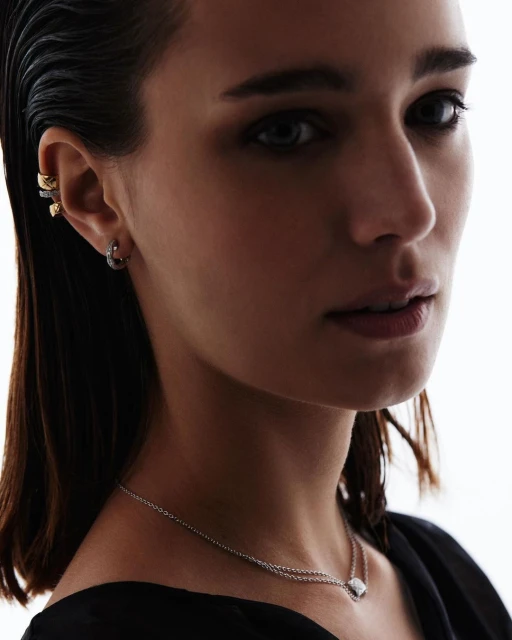 Close-up portrait of a woman with dark hair wearing earrings and a necklace against a bright white background, focusing on jewelry details.